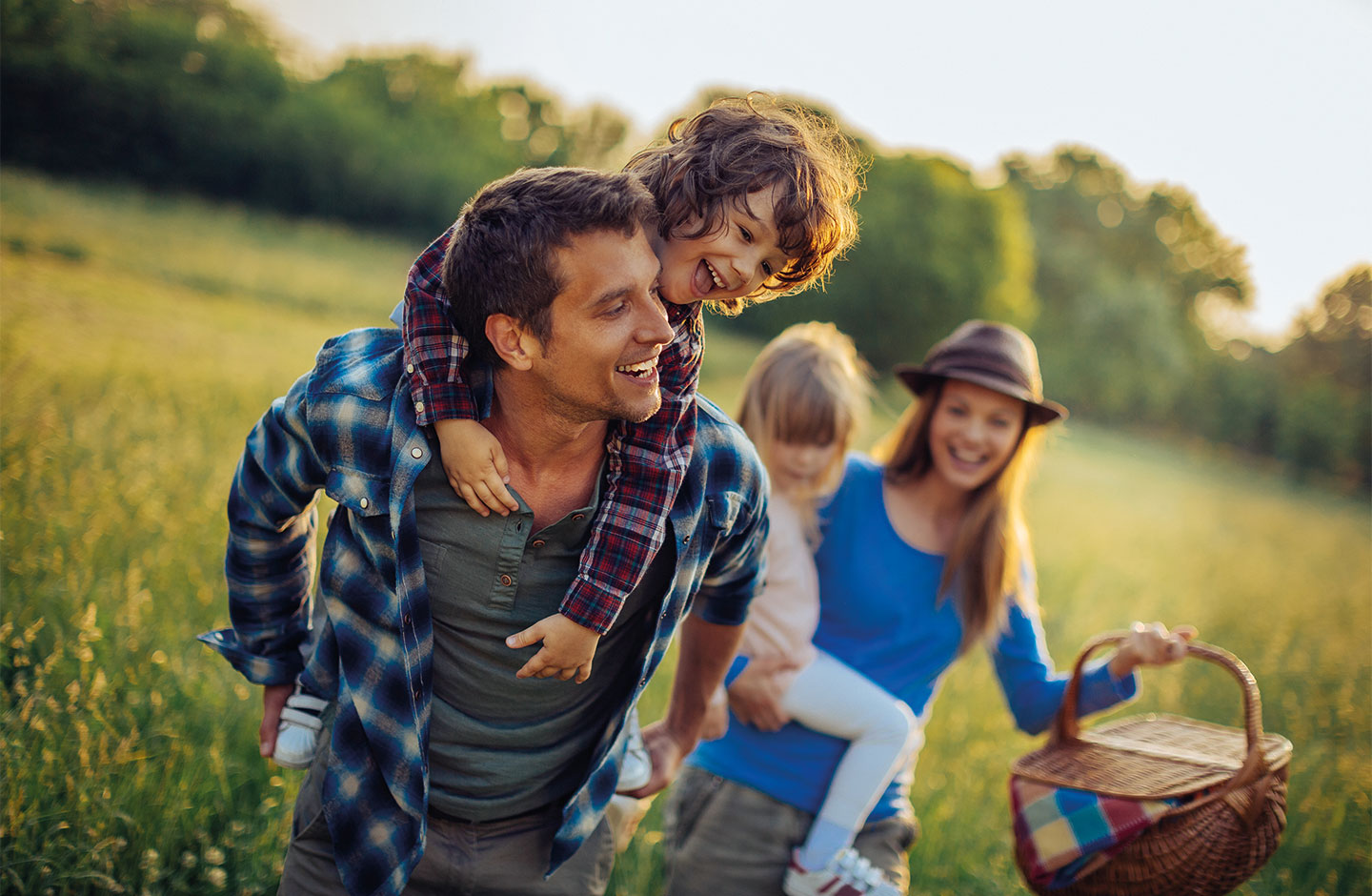 Junge Familie in der Natur. Mann trägt ein Kind am Rücken, Frau mit zweitem Kind im Hintergrund.