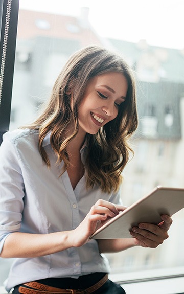 Eine junge Frau in Business-Bluse lehnt lächelnd an einem Fenster und tippt auf einem Tablet.
