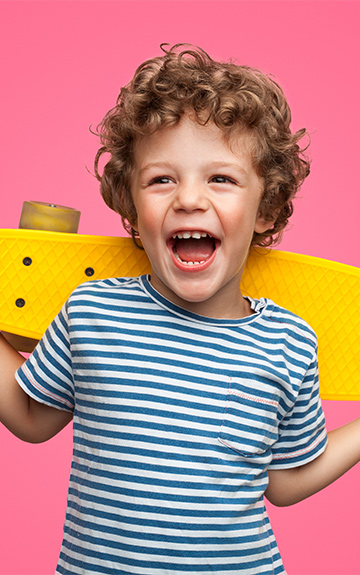 Ein kleiner Junge mit Locken lacht in die Kamera und hält ein gelbes Skateboard hinter seinem Rücken fest.