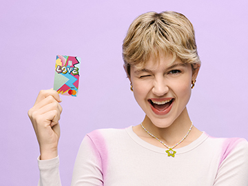 Eine junge Frau mit kurzen blonden Haaren hält eine Volksbank Debit Master Card in der Hand und zwinkert in die Kamera.