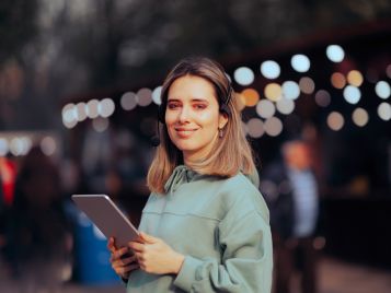 Eine Frau hält ein Tablet in der Hand und lächelt in die Kamera. Im Hintergrund ist verschwommen ein Großevent zu erahnen.