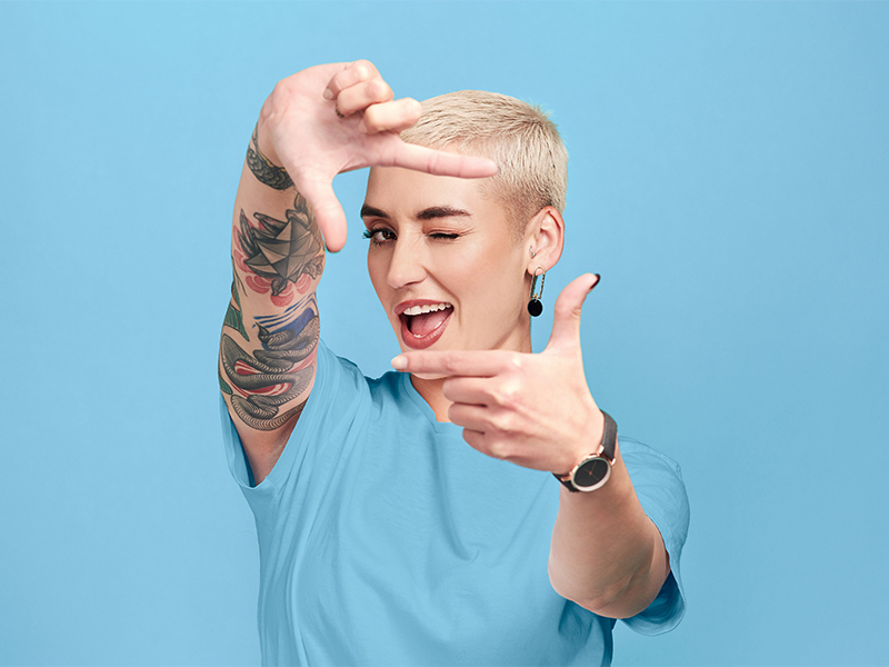 Eine Frau mit kurzem, blonden Haar steht vor einem hellblauen Hintergrund und formt mit ihrem Fingern ein Rechteck. Sie zwinkert in die Kamera. 