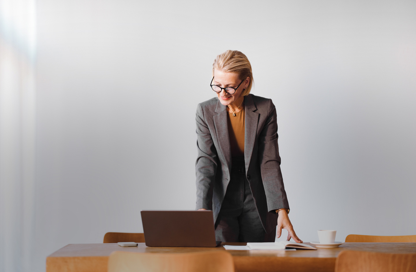 Eine ältere Frau im Blazer stützt sich mit den Händen auf einen Tisch und blickt lächelnd auf einen Laptop