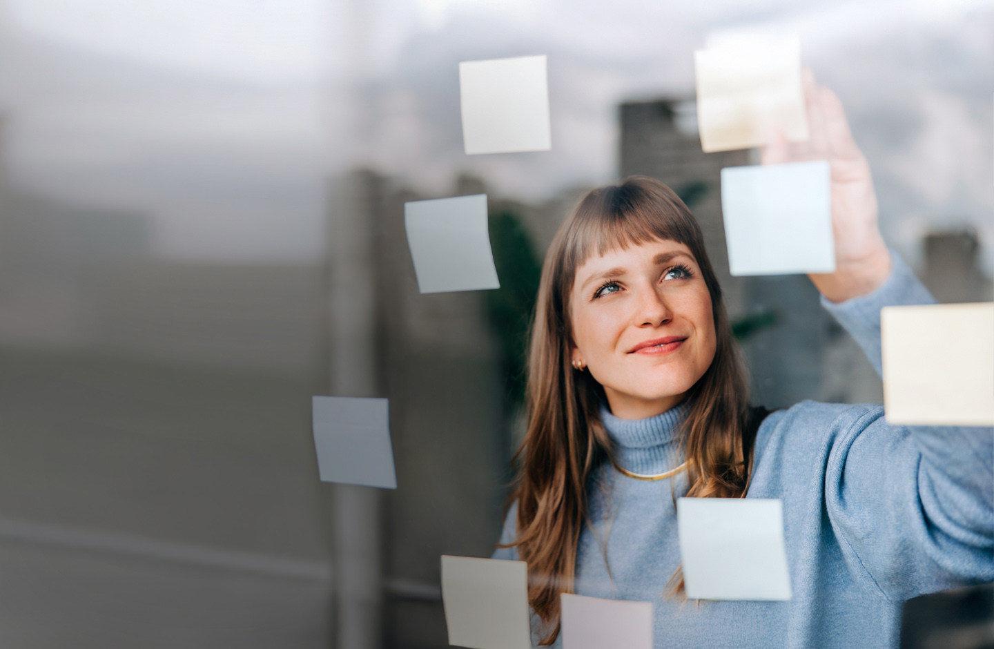 Eine junge Frau mit blondem Haar klebt lächelnd bunte Post-its an eine Fensterscheibe.
