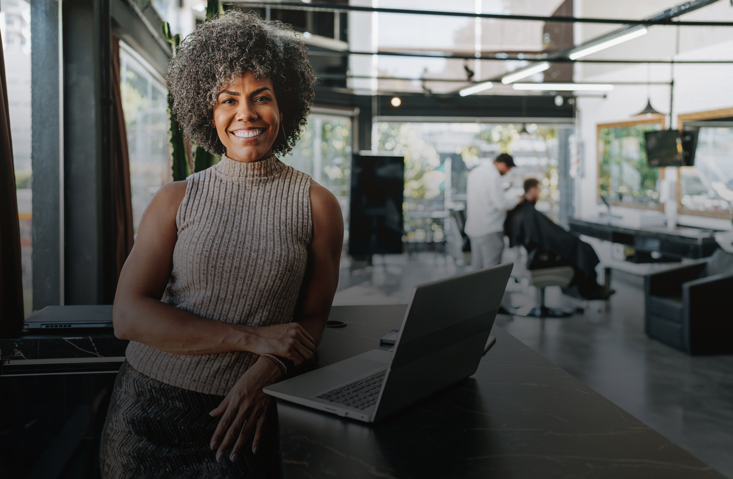 Eine Frau im Business-Dress mit Afro steht in einem Büro und lehnt sich lässig an einen Tisch auf dem ein Laptop steht.