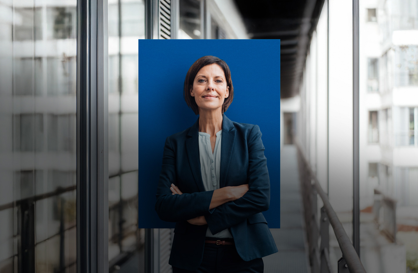 Eine Frau im Business Dress steht in einem Büro. Hinter ihr befindet sich ein blaues Quadrat.