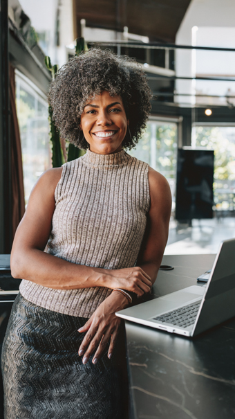 Eine im Business-Dress gekleidete Frau mit Afro lehnt an einem Tisch auf dem ein Laptop steht