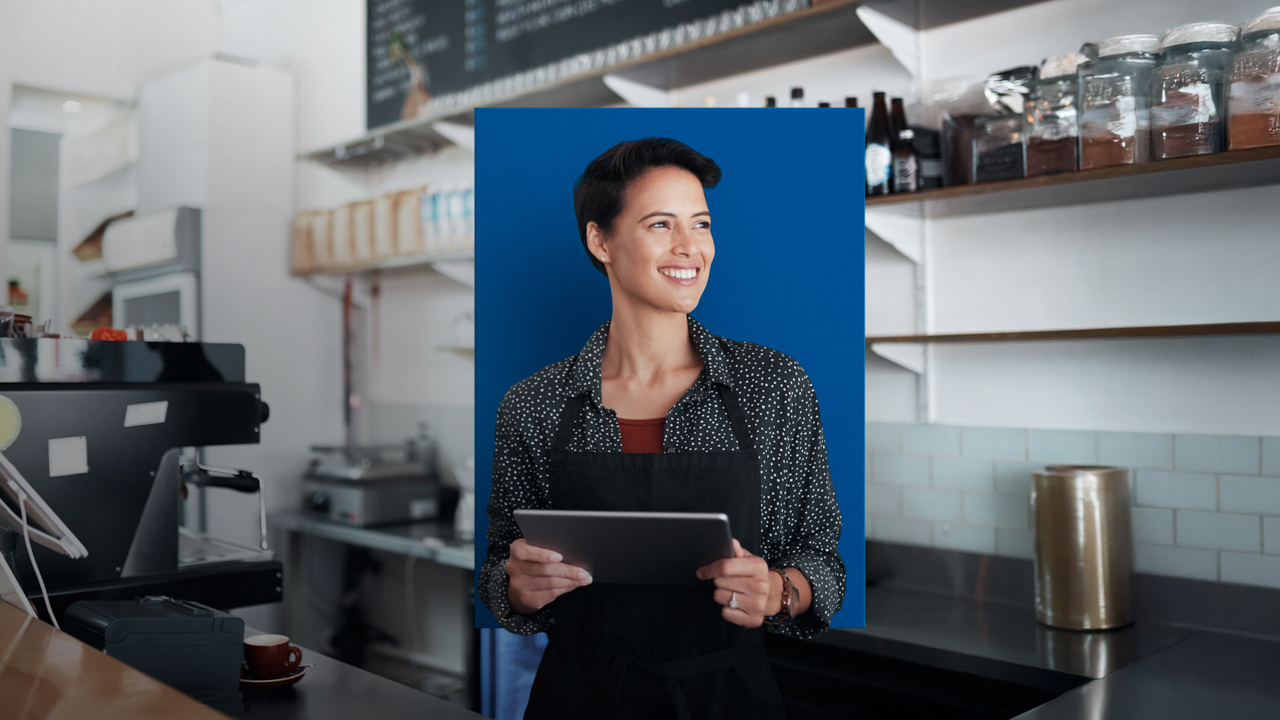 Eine Frau steht in einer Kaffeeküche mit Schürze und hält lachend ein Tablet in der Hand.