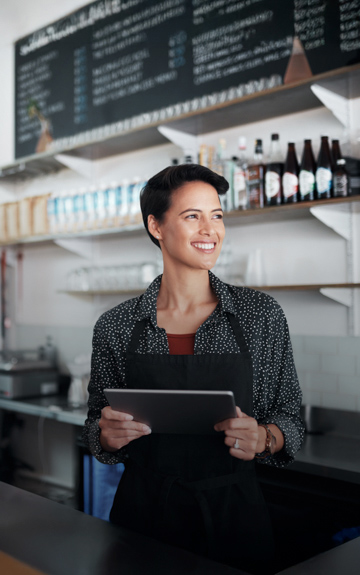 Eine junge Frau steht lachend mit Tablet in der Hand in einem Café und blickt in die Ferne.