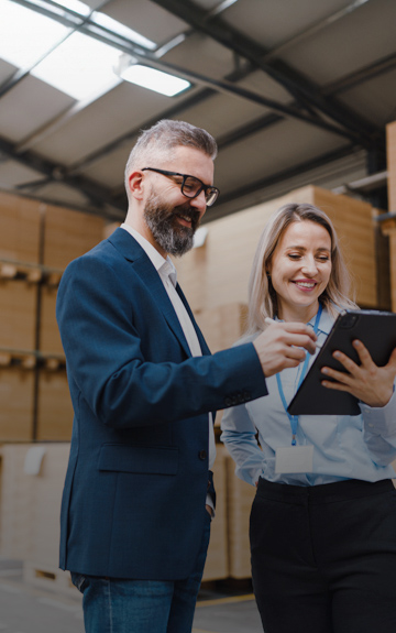 Ein Mann im Anzug und eine Frau in Business-Bluse stehen in einer Lagerhalle und blicken lachend auf ein Tablet.