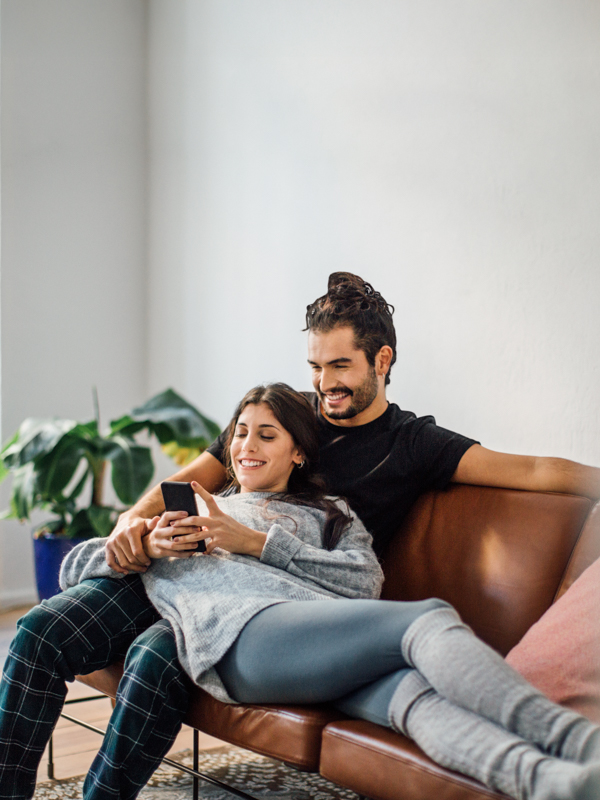 Eine Frau liegt in den Armen eines Mannes auf einer Couch. Gemeinsam blicken Sie auf ein Handy.