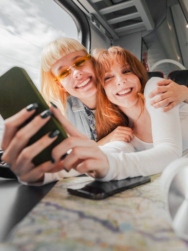 Zwei junge Frauen sitzen in einem Zug. Sie umarmen sich und blicken gemeinsam auf ein Mobiltelefon.