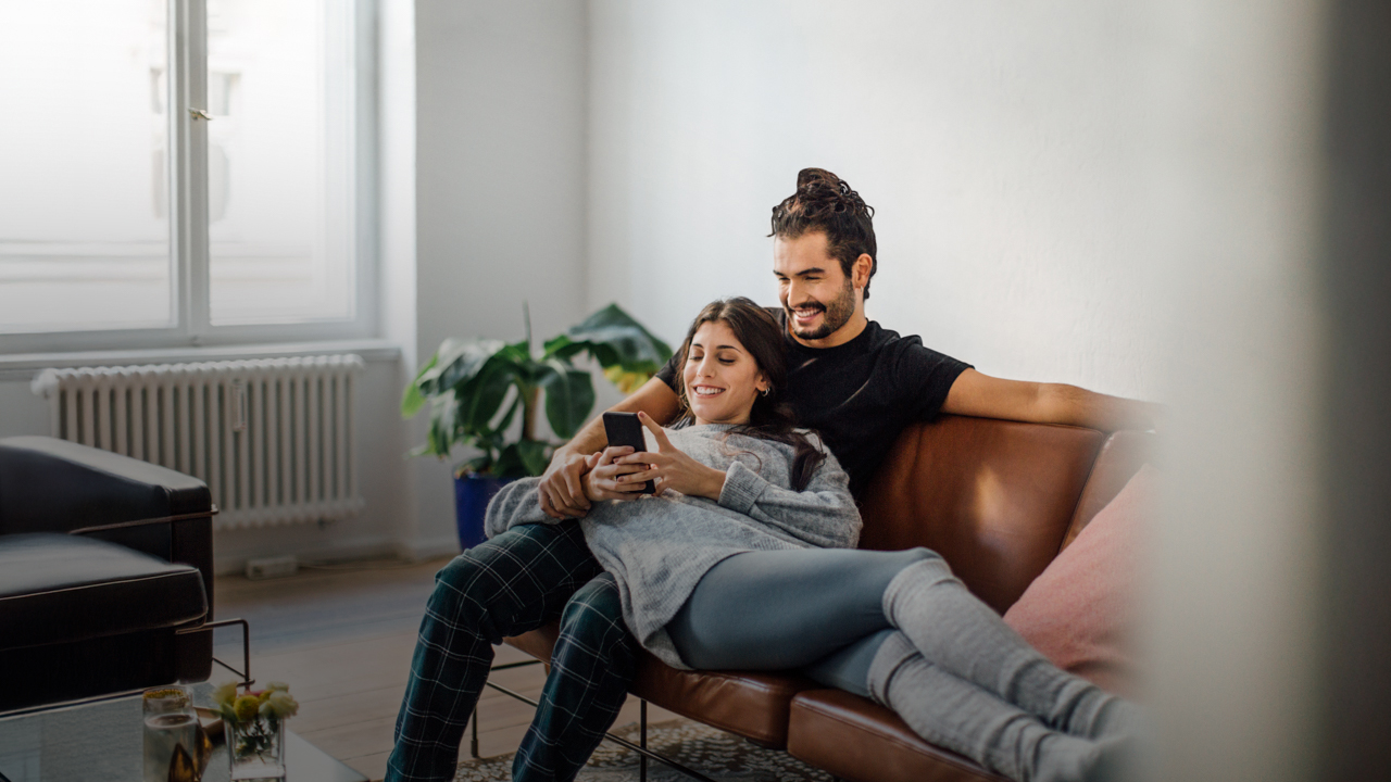 Ein Mann sitzt auf einer Couch, eine Frau liegt in seinem Schoß. Sie blicken gemeinsam auf ein Mobiltelefon.