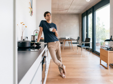 Ein Mann lehnt an einem Tresen in einer Küche mit Kaffeetasse in der Hand.