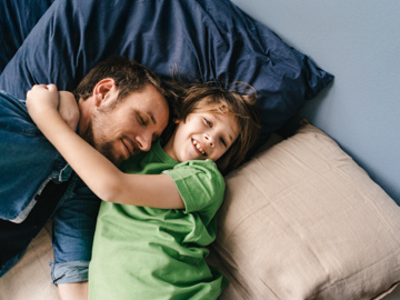 Ein Vater und sein Sohn liegen auf einer Couch. Der Sohn umarmt den Vater.