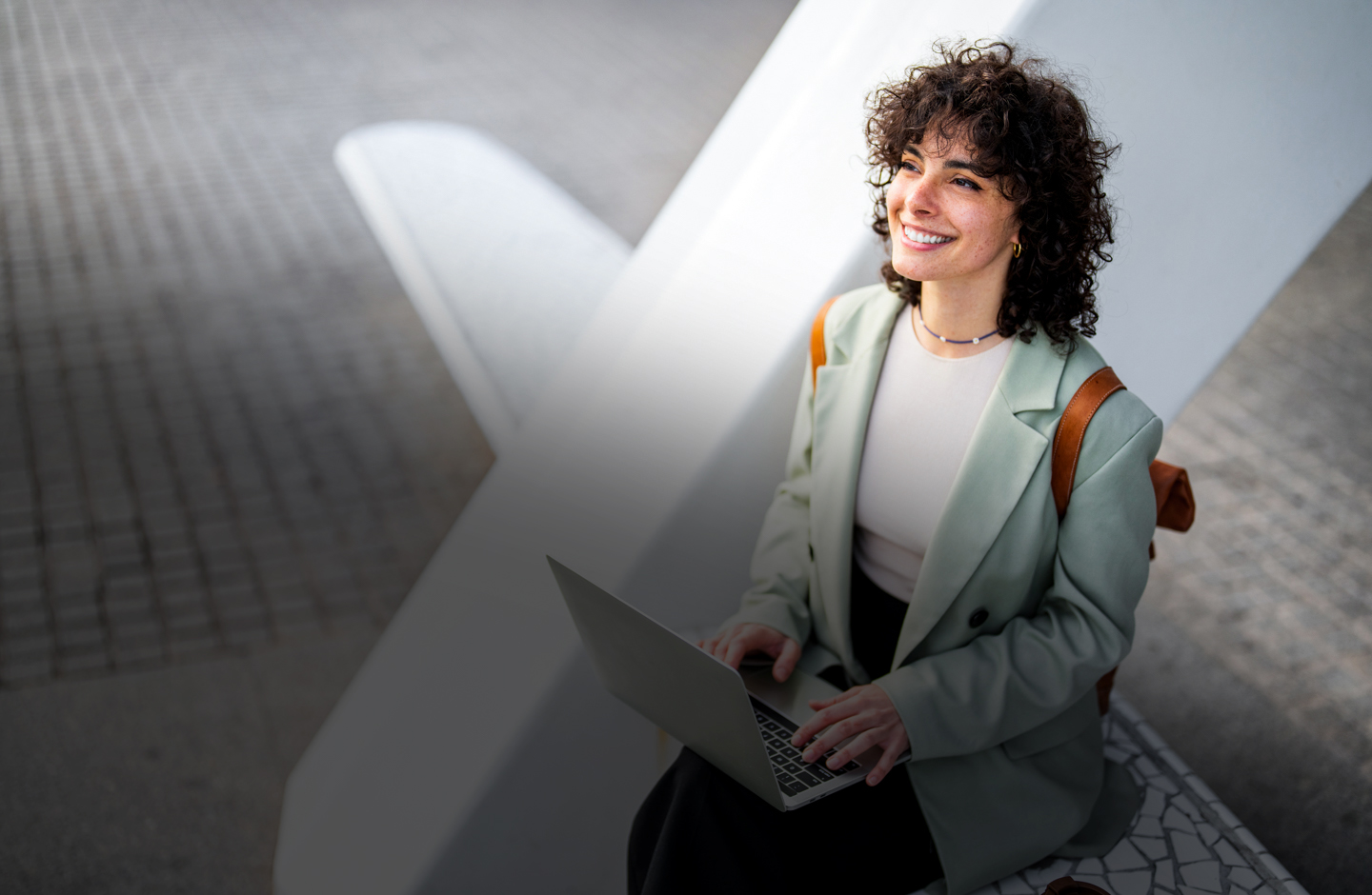 Eien junge Dame sitzt draußen auf einer weißen Bank mit Laptop auf dem Schoß und schaut nach oben.